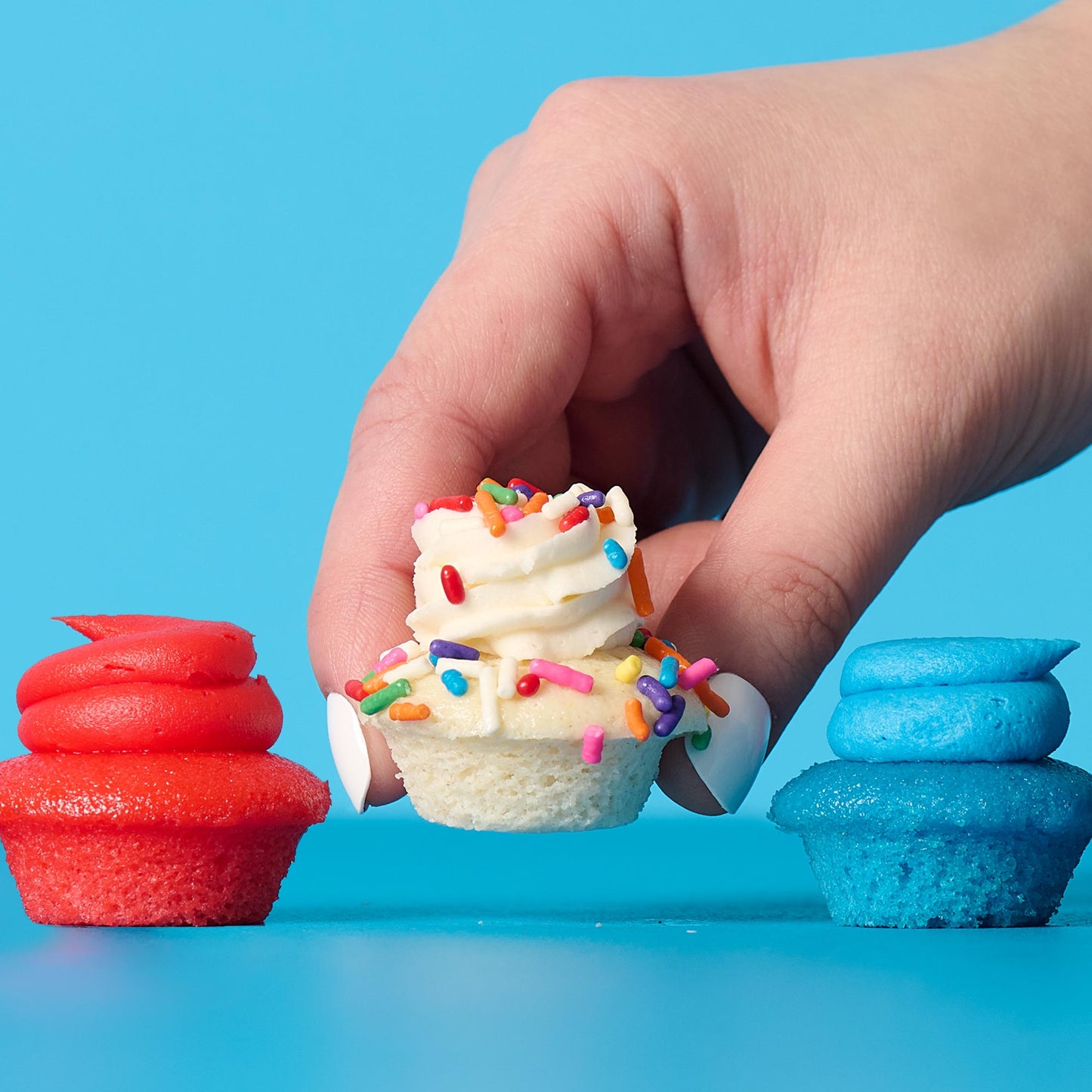 Hand holding a small cupcake with colorful sprinkles between two cupcakes, one red and one blue, on a blue background.